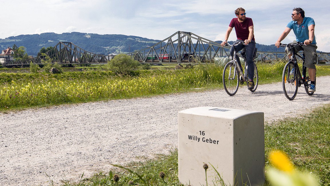 Zwei Männer fahren mit dem Fahrrad über einen Weg, im Vordergrund des Bildes ist ein Gedenkstein mit der Aufschrift "Willy Geber" zu sehen
