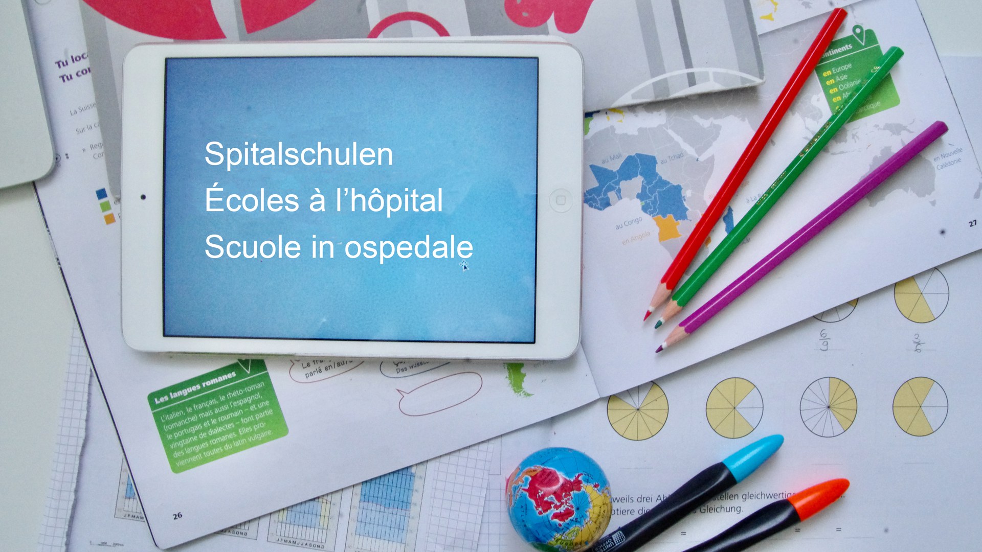 Une tablette posée sur une table, entourée de matériel scolaire et sur l’écran de laquelle on peut lire «Écoles à l’hôpital»