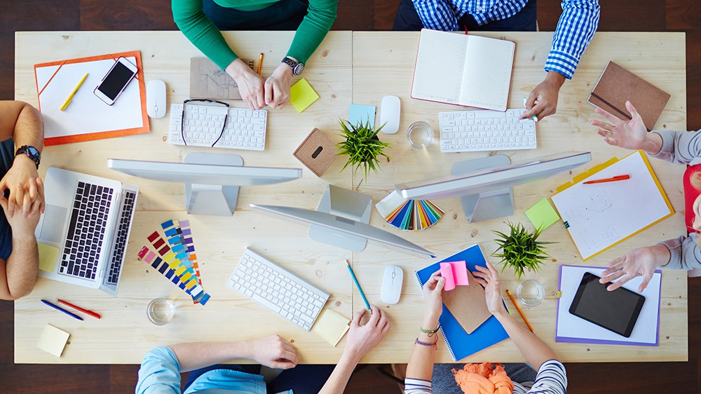 Vue d'en haut sur une table sur laquelle sont posés des ordinateurs, des agendas, des feuilles de notes et des stylos